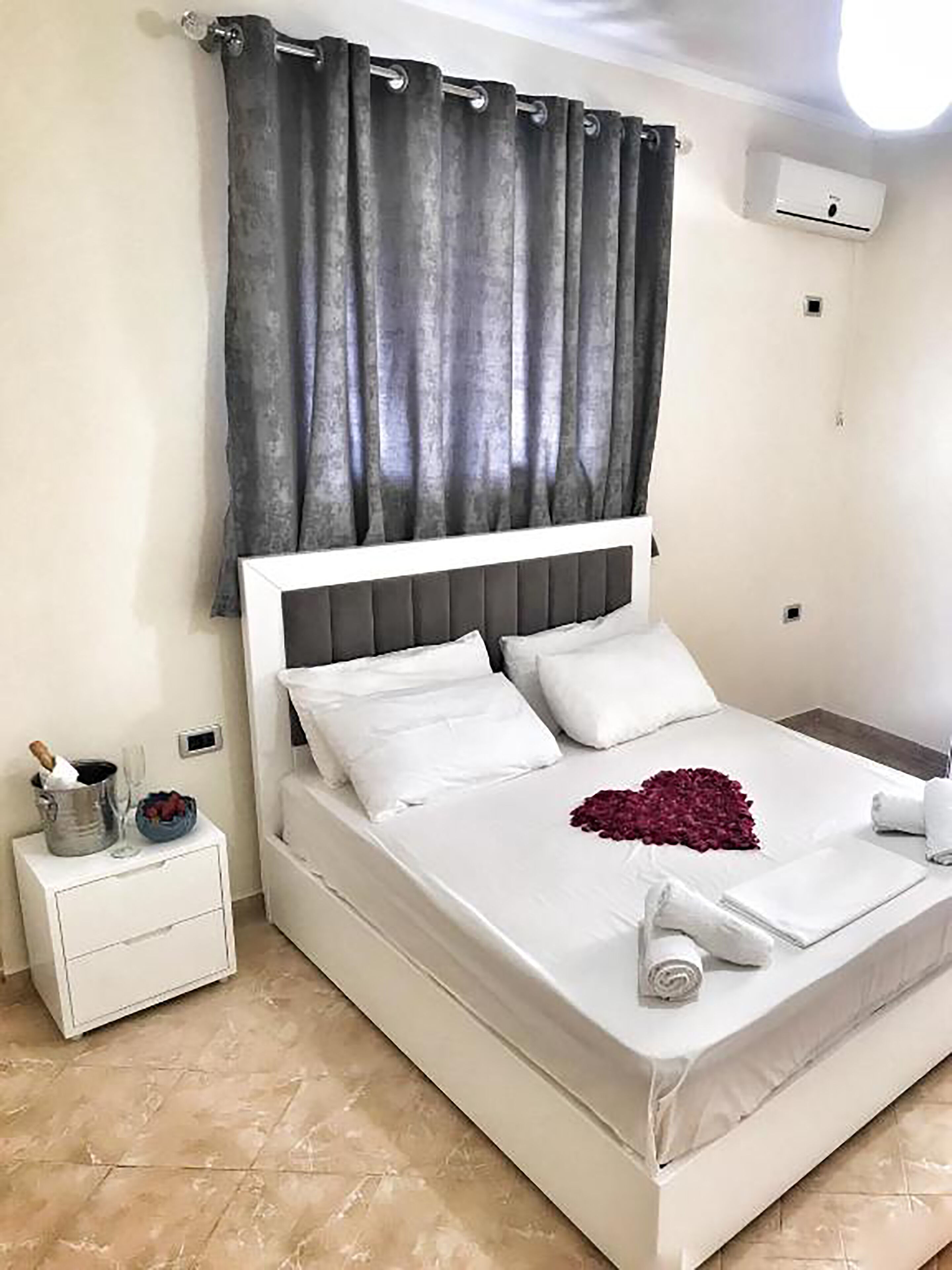 A neatly made bed with white linens and a heart-shaped decoration, a small bedside table with a champagne bottle and glasses, and a grey curtain.