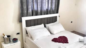 A neatly made bed with white linens and a heart-shaped decoration, a small bedside table with a champagne bottle and glasses, and a grey curtain.
