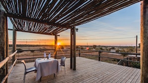 Une terrasse en bois avec une table dressée pour deux personnes, offrant une vue sur un paysage désertique ponctué de lignes électriques et illuminé par le coucher du soleil.