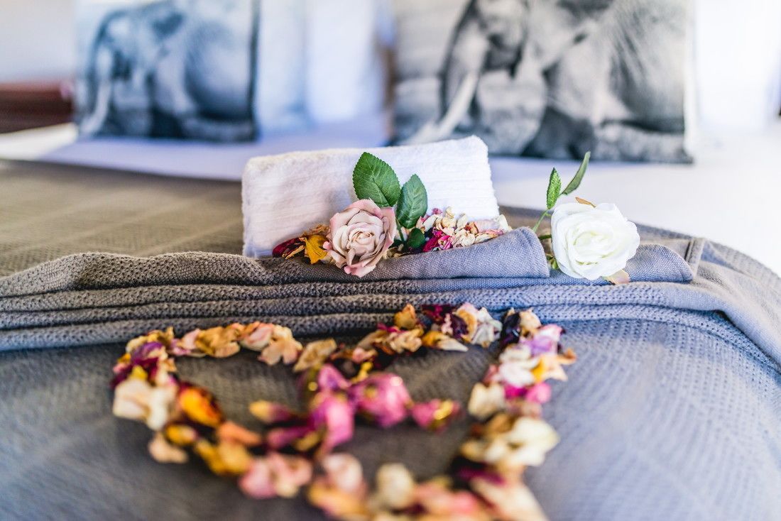 Ein Bett mit grauer Decke, einem weißen Kopfkissen und einem beigen Handtuch. Auf der Decke liegen getrocknete Blumen und eine rosa Rose.