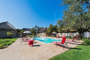 A pool area with red lounge chairs and umbrellas, surrounded by buildings with thatched roofs.