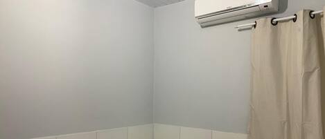 Una habitación pequeña con una cama individual, cabecera blanca, manta marrón y almohada blanca. Unidad de aire acondicionado montada en la pared sobre la cama.