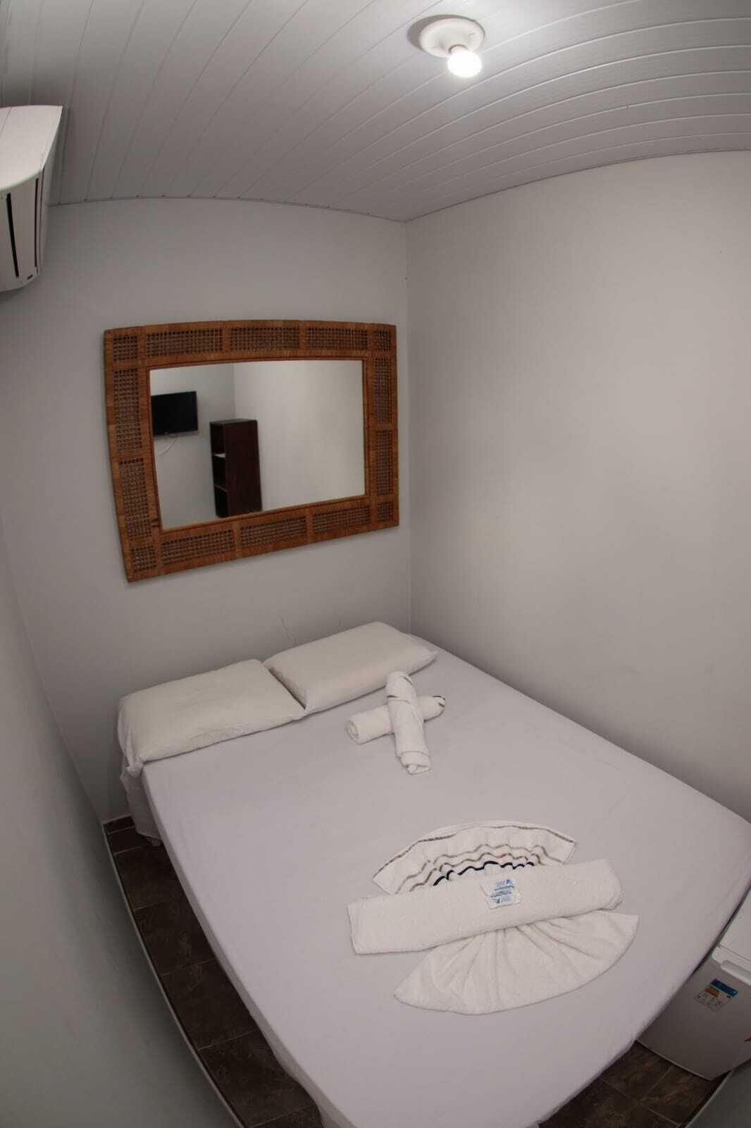 Una habitación pequeña con una cama individual, una cama blanca sencilla, un espejo con marco de madera y una pared blanca.