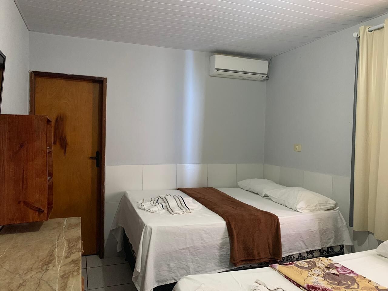 Una habitación pequeña y limpia con dos camas individuales, un armario de madera y un equipo de aire acondicionado en la pared.