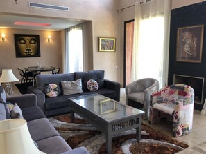 A modern living room with a sofa, armchair, and coffee table. There is a dining area with a table and chairs, and a fireplace. - Villa les Jardins d'Atlas (Marrakech)