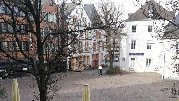 A cobblestone courtyard with outdoor seating, a white building with a blue sign, and a tree in the foreground. - Ricks City Hotel 2 (Trier)