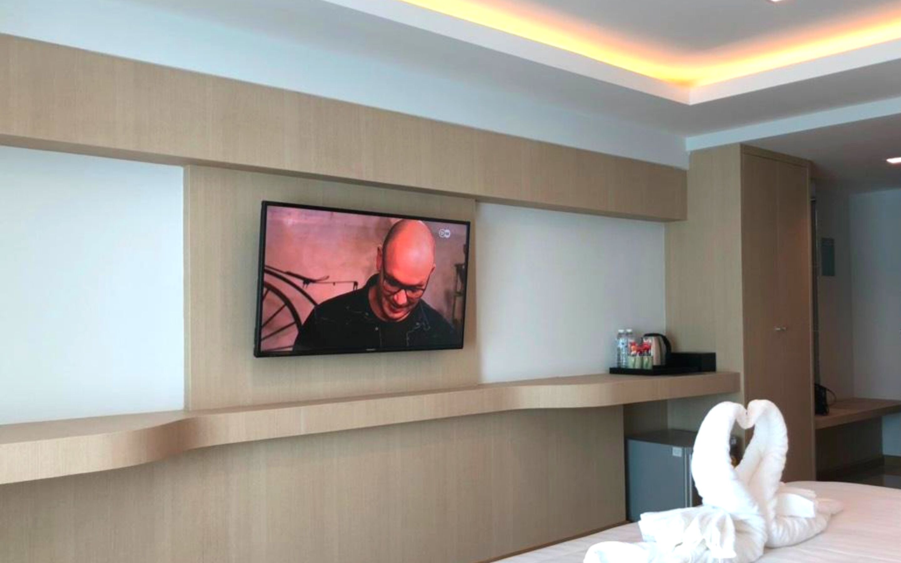 A hotel room with a mounted TV on the wall, a bed with a white swan-shaped pillow, and a minibar with beverages.