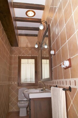 A bathroom with a toilet, sink, and mirror, featuring a tiled wall and wooden ceiling beams. - Casa Rural Aingeru (Valle de Carranza)