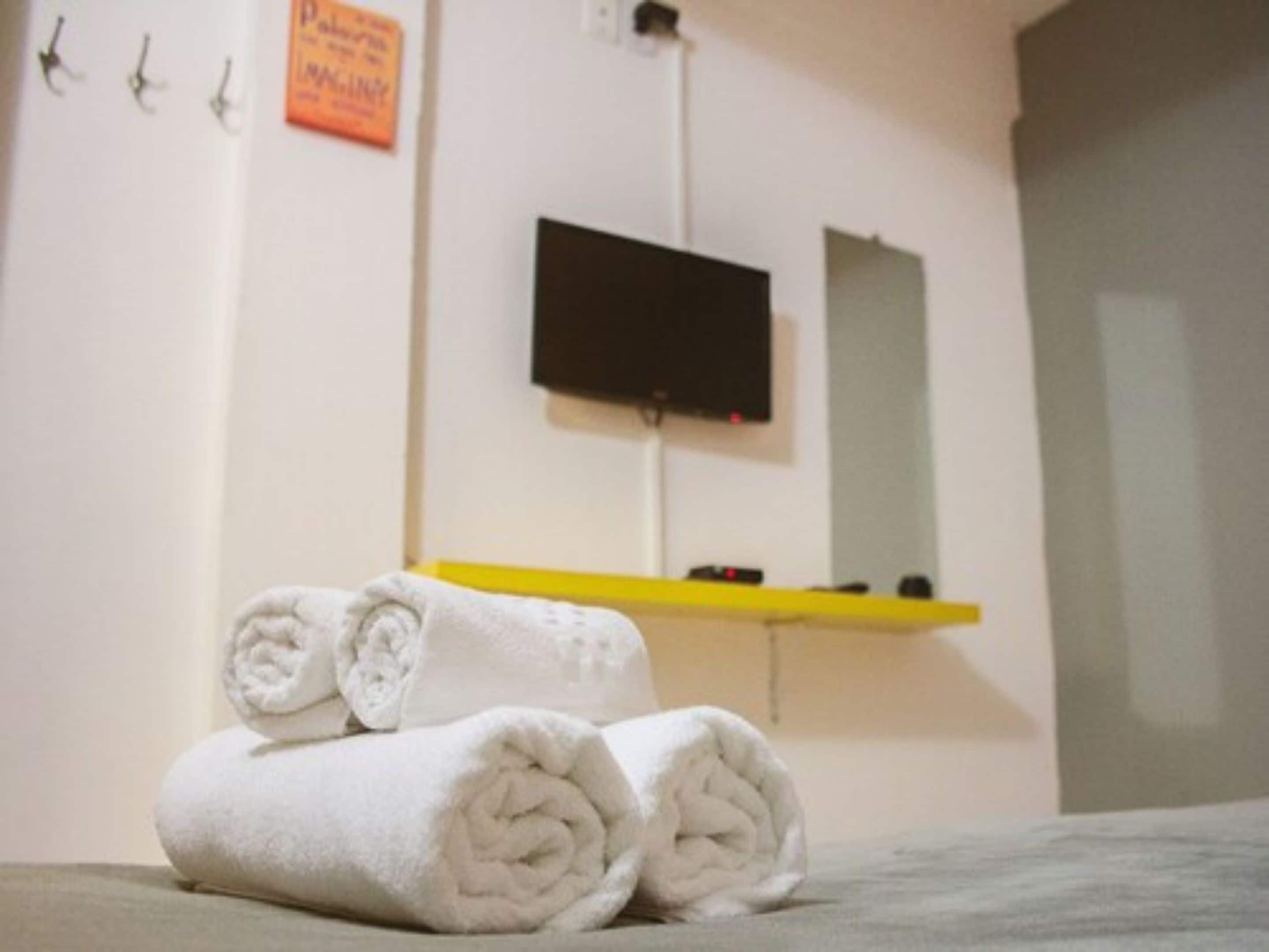 A bathroom with a white towel rolled up on the floor, a yellow shelf with a black object on it, and a flat-screen TV mounted on the wall.