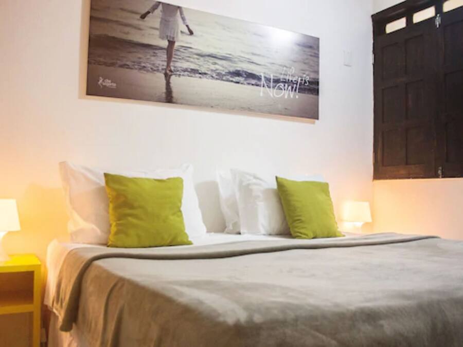 Un dormitorio con cama, almohadas, una mesita de noche, un cuadro en la pared y una puerta.