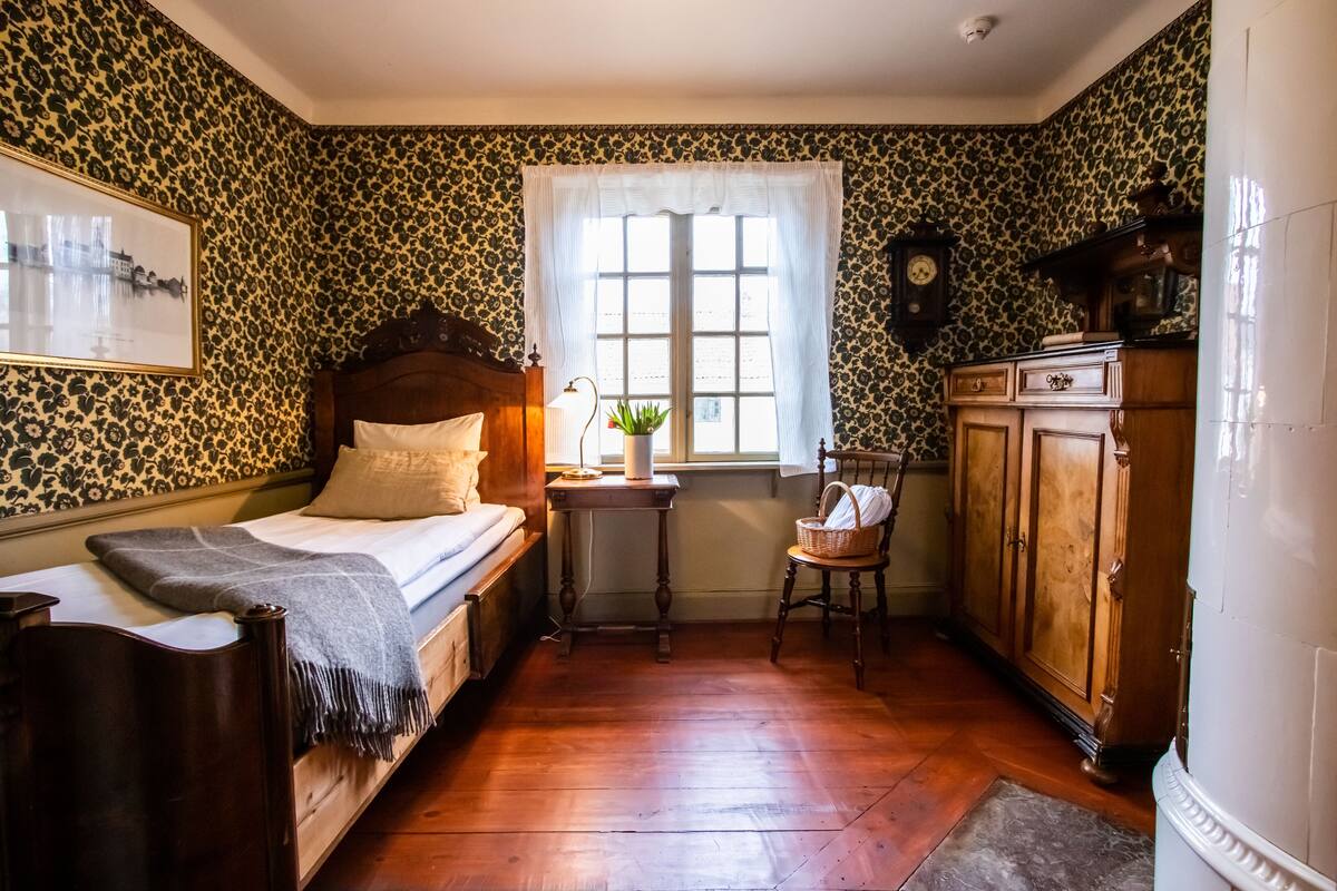 Un dormitorio con una cama de madera, una mesita de noche, una ventana con cortinas, un armario de madera y un reloj de pared.
