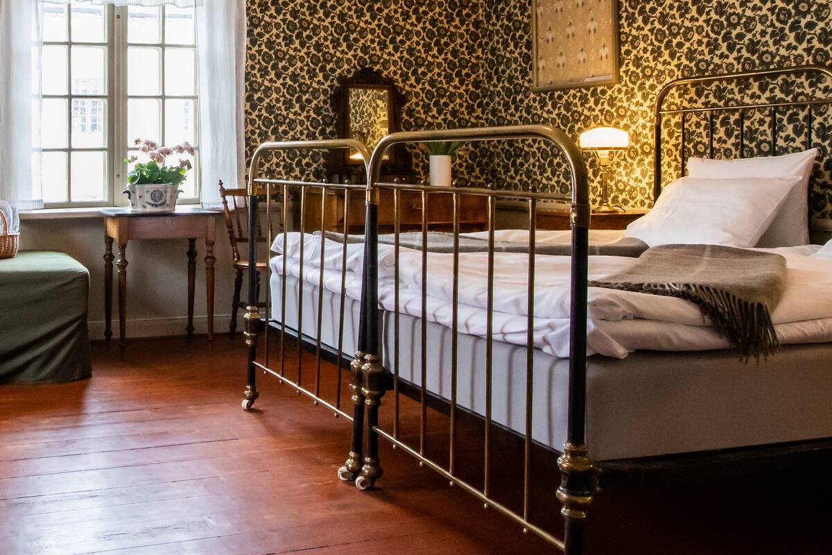 Un dormitorio con estructura metálica en la cama, una mesita de noche, una lámpara y una ventana con cortinas.