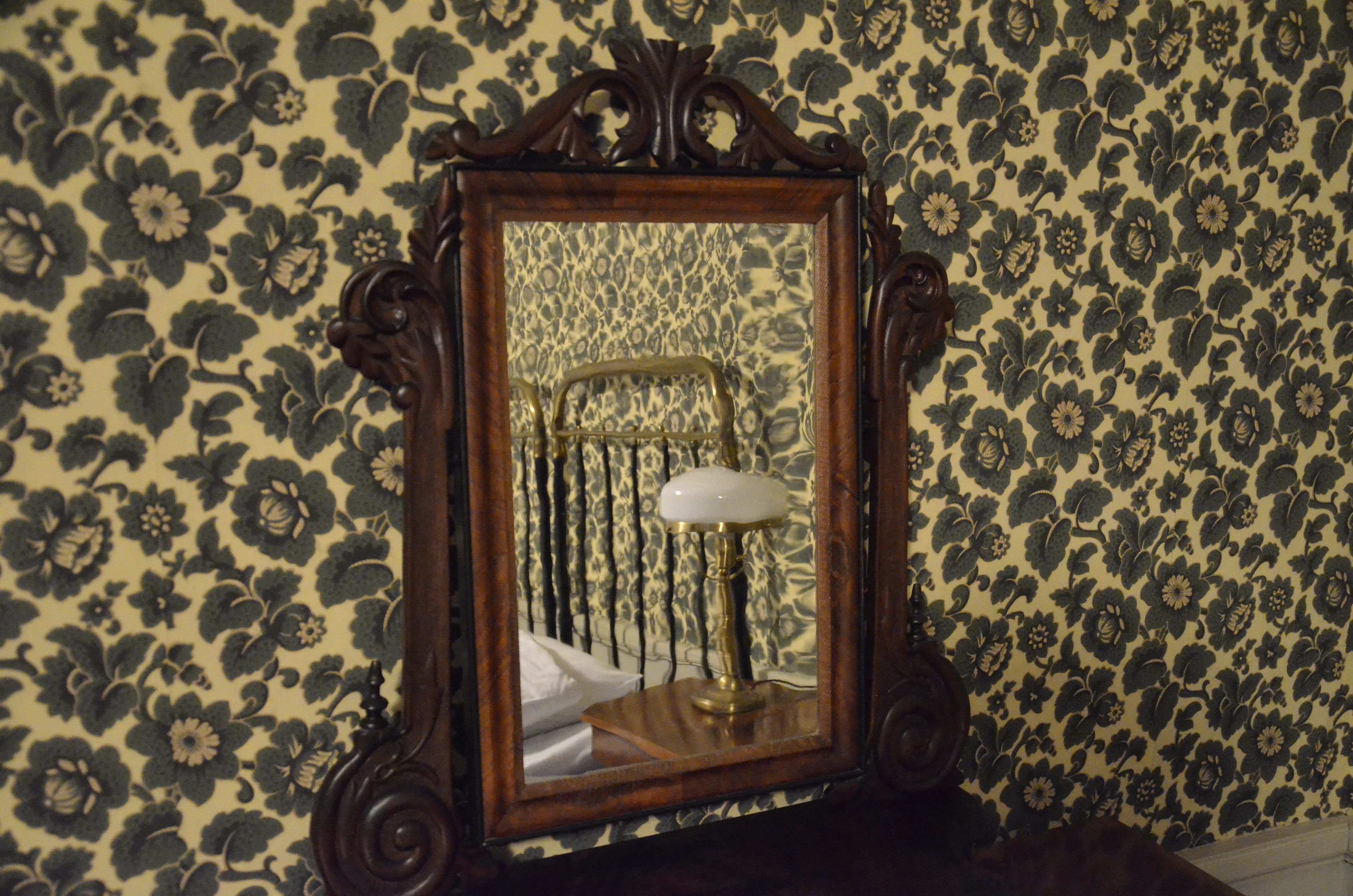 Un espejo con un marco de madera decorado refleja una cama con un cojín blanco y una lámpara de latón.