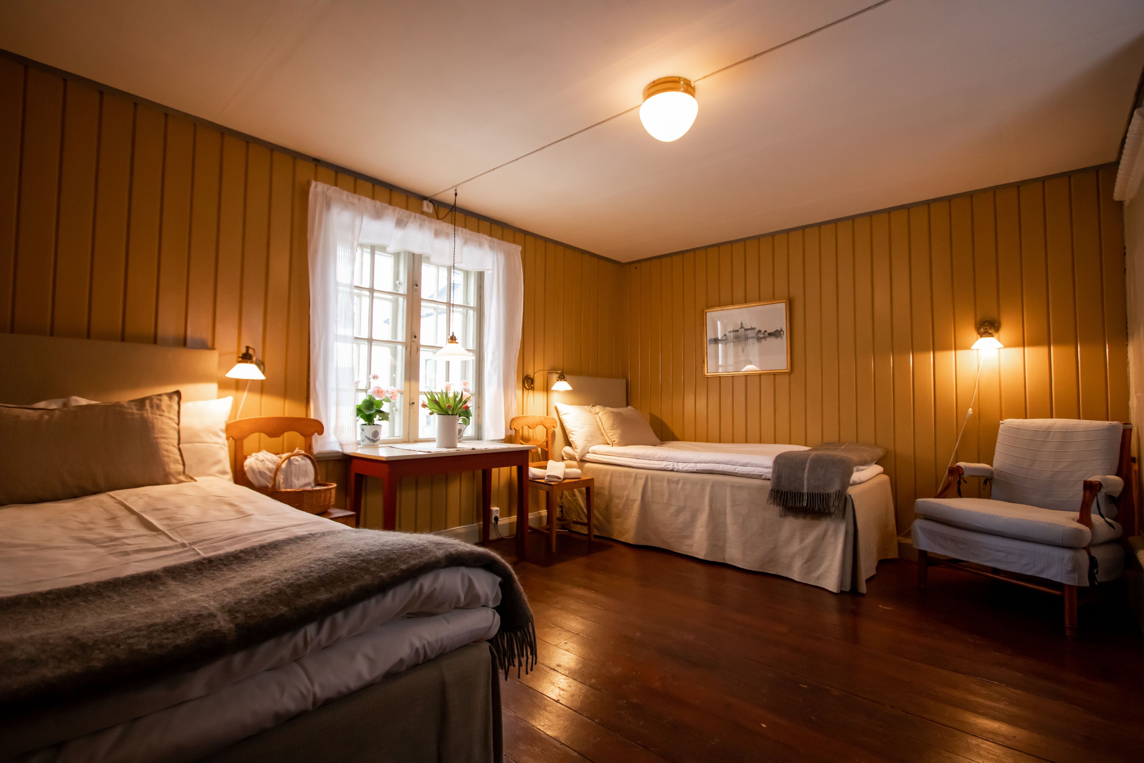 Een hotelkamer met twee bedden, een kleintje tafeltje met een plant, een stoel en een wandlamp.