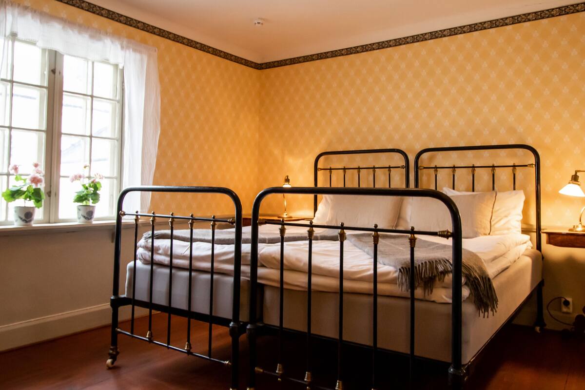 Un dormitorio con estructura metálica para la cama, una mesita de noche con lámpara y una ventana con cortinas.