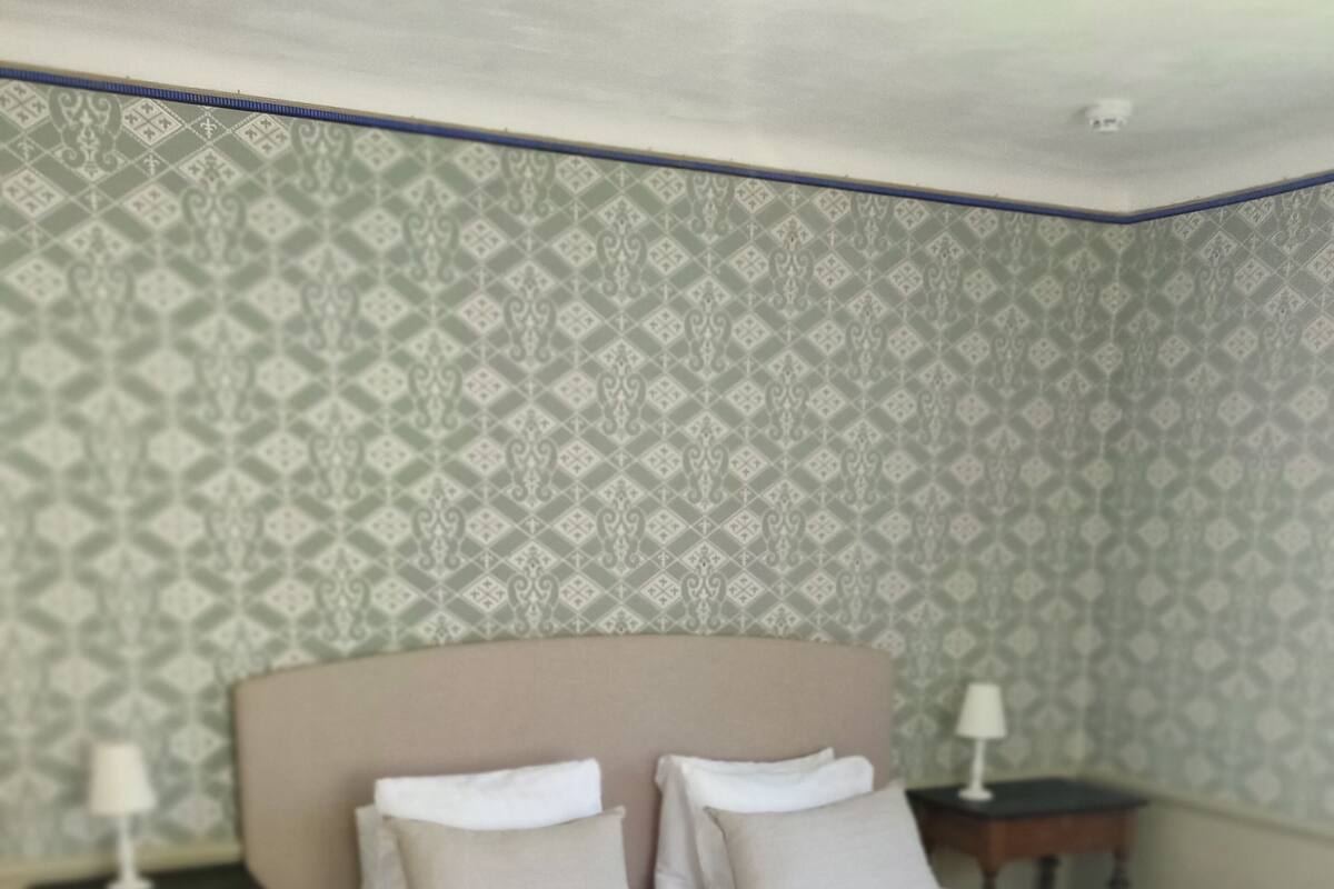 Una cama doble con una colcha gris, dos mesitas de noche con lámparas y un papel pintado estampado.