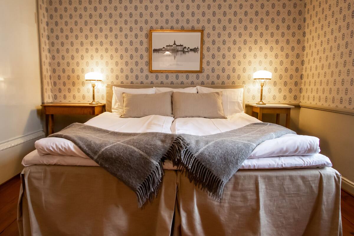 Una cama bien hecha con una colcha y almohadas, dos lámparas de noche y un cuadro en la pared.