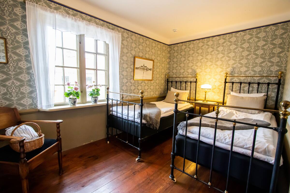 Una habitación con dos camas, una ventana con vistas, una silla con una cesta y papel tapiz estampado.