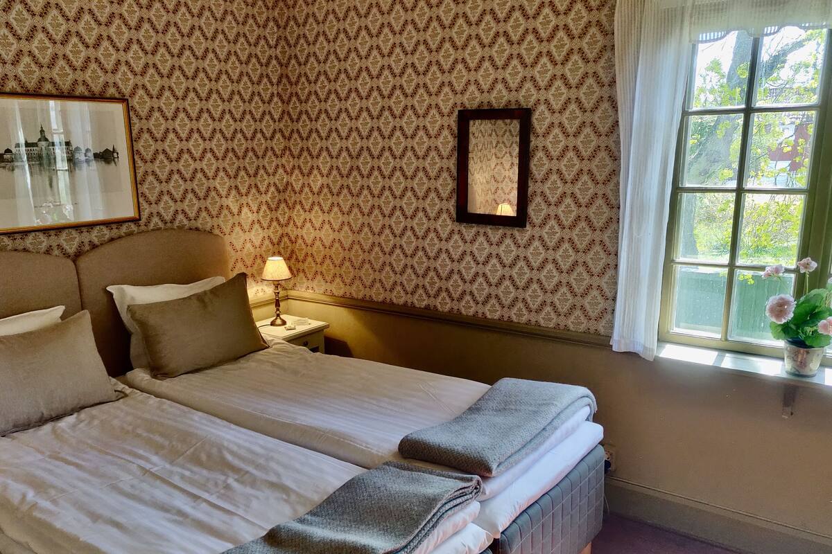Una cama doble bien hecha con almohadas, una mesita de noche con lámpara, un cuadro enmarcado en la pared y una ventana con vistas a la vegetación.