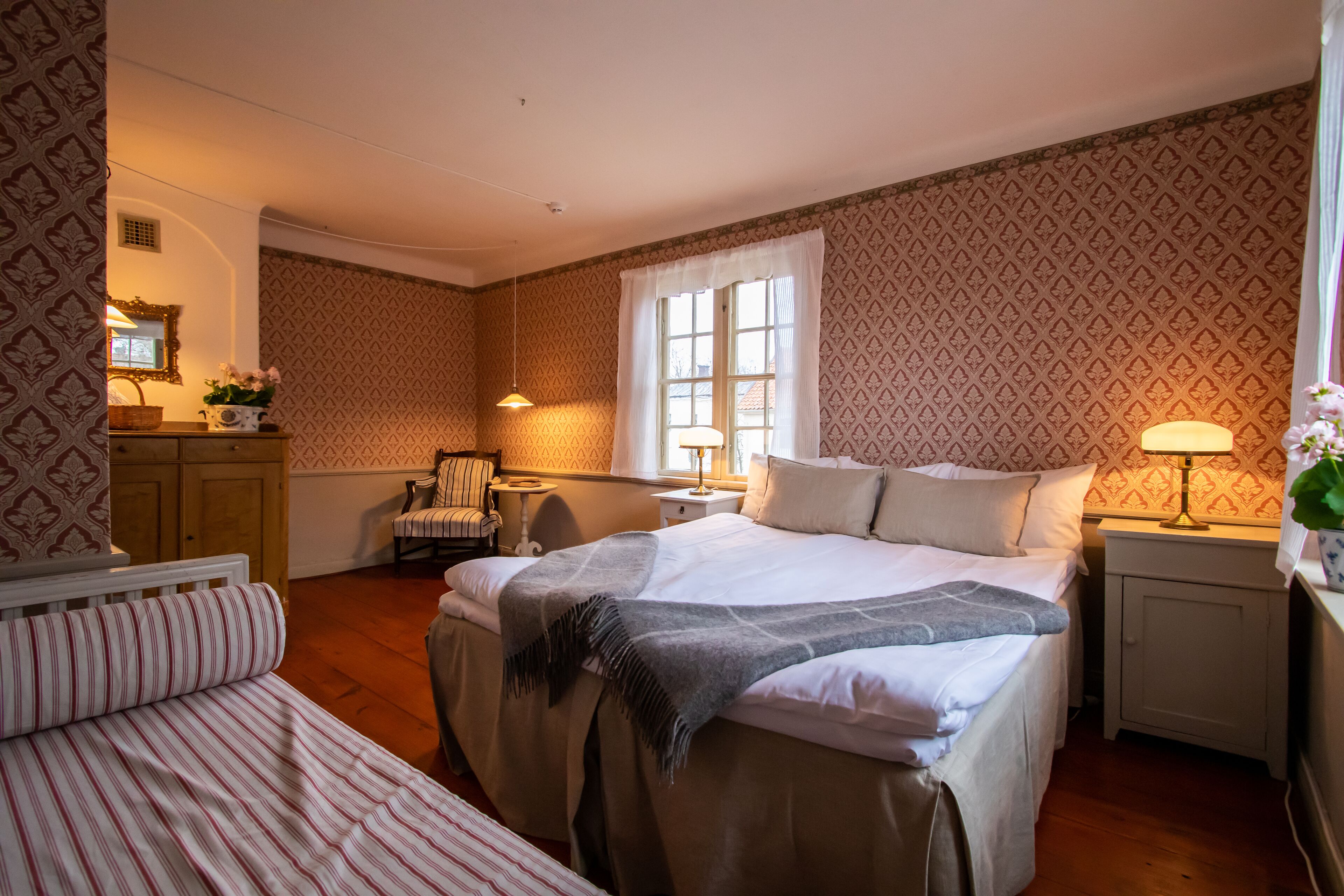 A bedroom with a large bed, a smaller bed, a chair, a desk, and a window with curtains.
