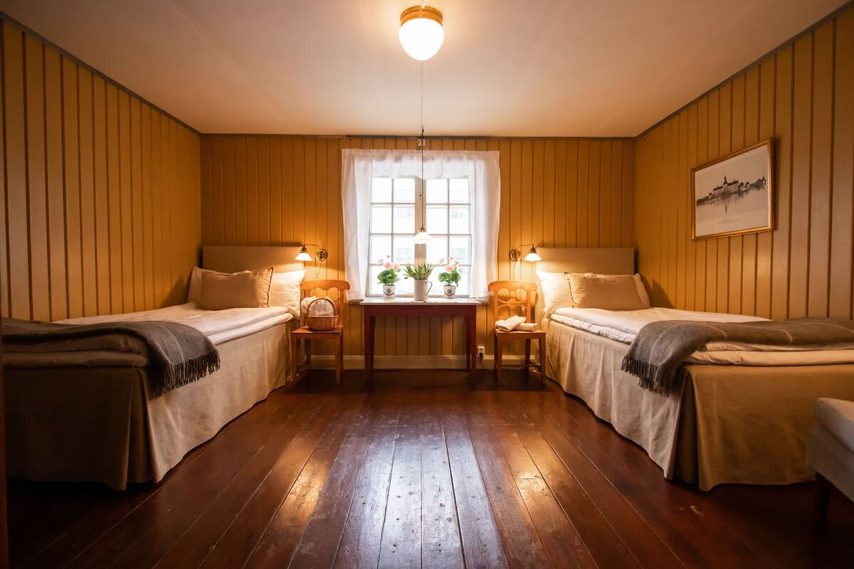 Una habitación con dos camas individuales, paneles de madera, un ventanal con vistas y una pequeña mesa con un jarrón y flores.
