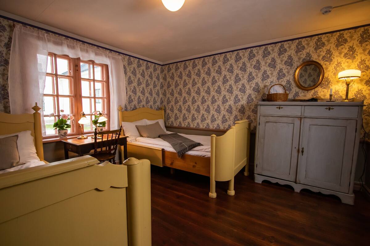 Un dormitorio con dos camas individuales, un aparador de madera, un escritorio con silla, una ventana con cortinas y un papel tapiz estampado.