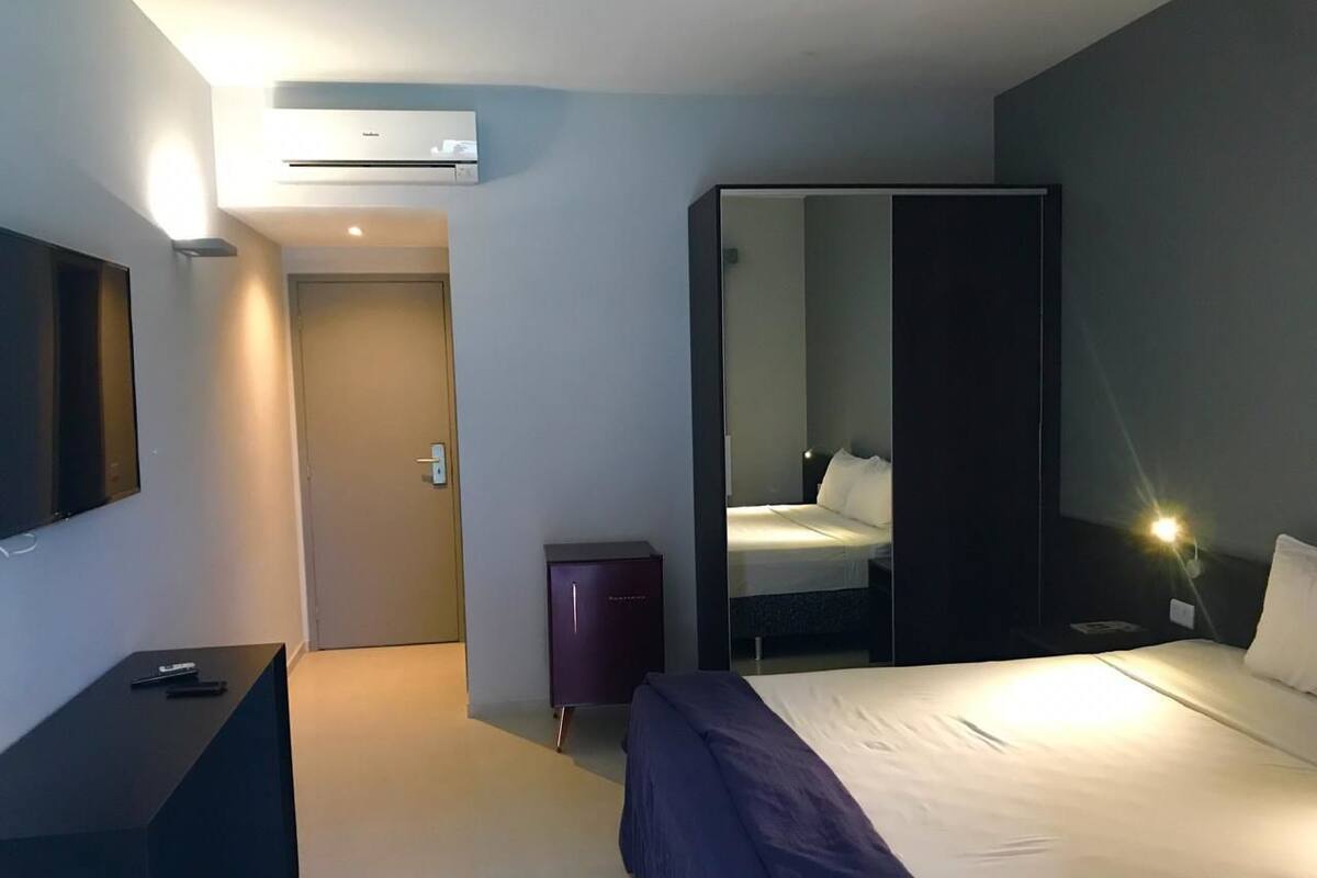 Quarto de hotel com cama, escrivaninha, guarda-roupa, espelho e ar condicionado.