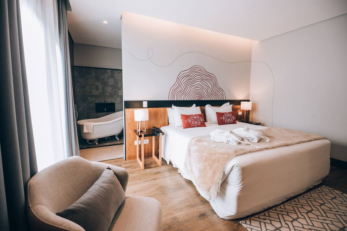 Una habitación de hotel moderna con una cama grande, lámparas de noche, una mesita de noche de madera, una silla y un baño con bañera.