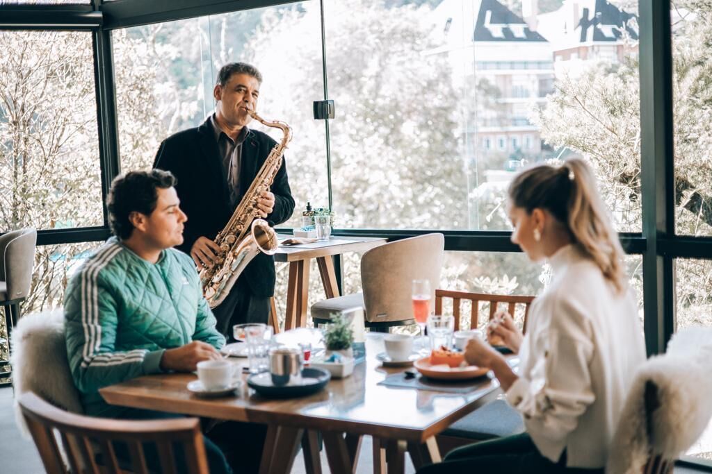 Un comedor con una mesa dispuesta para tres personas, un saxofonista tocando y vistas a árboles y edificios a través de amplias ventanas.