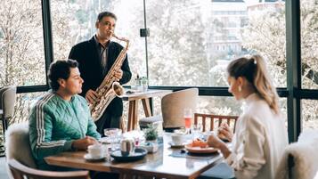 Un comedor con una mesa dispuesta para tres personas, un saxofonista tocando y vistas a árboles y edificios a través de amplias ventanas.