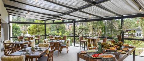 A spacious outdoor dining area with multiple wooden tables and chairs, surrounded by large windows offering a view of greenery and buildings.