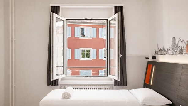 A hotel room with a bed, a window with curtains, a radiator, and a wall with a cityscape mural. - easyHotel Zürich City Limmatplatz (Zürich)