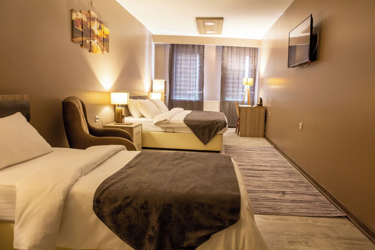Ein Hotelzimmer mit zwei Betten, einem Fernseher, einem Schreibtisch mit Lampe sowie einem Fenster mit Vorhängen.