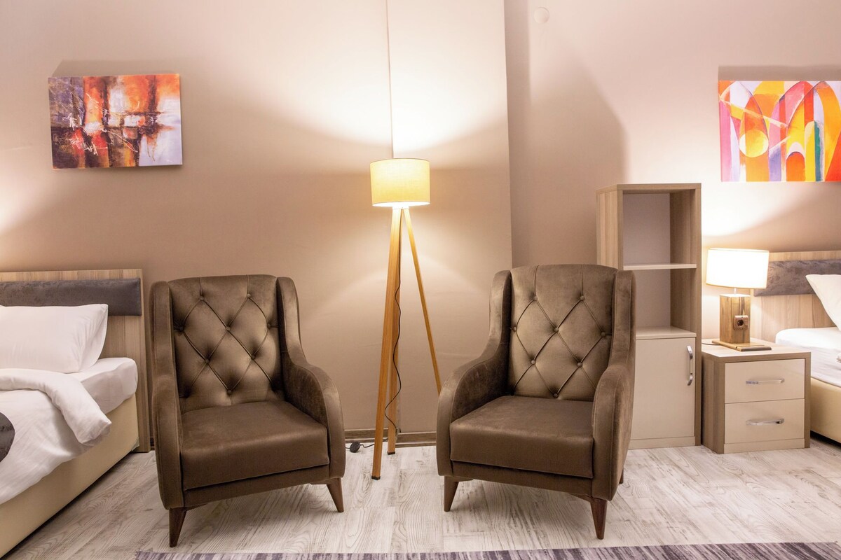 A hotel room with two tufted armchairs, a bedside table with a lamp, a bed, a nightstand, and abstract paintings on the walls.
