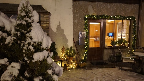 Un espace extérieur enneigé, avec un arbre recouvert de neige, une porte en bois ornée d’une couronne de Noël et un sapin à l’intérieur.