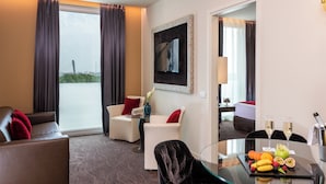 Habitación de hotel con sofá, dos sillones, una mesa de vidrio con fruta y copas de champán, y un televisor en la pared.