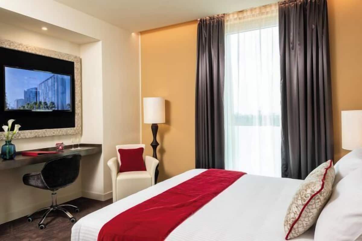 Una habitación de hotel con cama, escritorio, silla, televisor y ventana con cortinas.