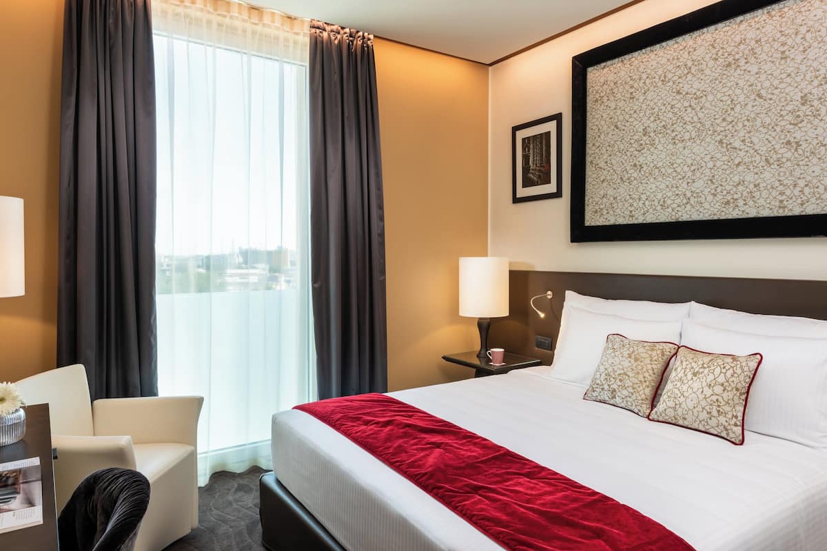 Habitación de hotel con una cama grande, un escritorio con silla, una ventana con cortinas, una lámpara de pared y una obra de arte enmarcada.
