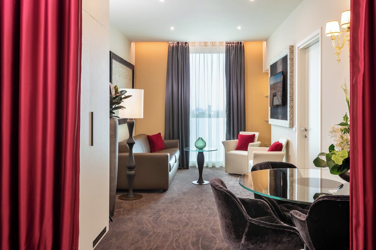 Habitación de hotel con sofá, sillones, una mesa de vidrio, lámpara y vistas a la ciudad a través de grandes ventanales.