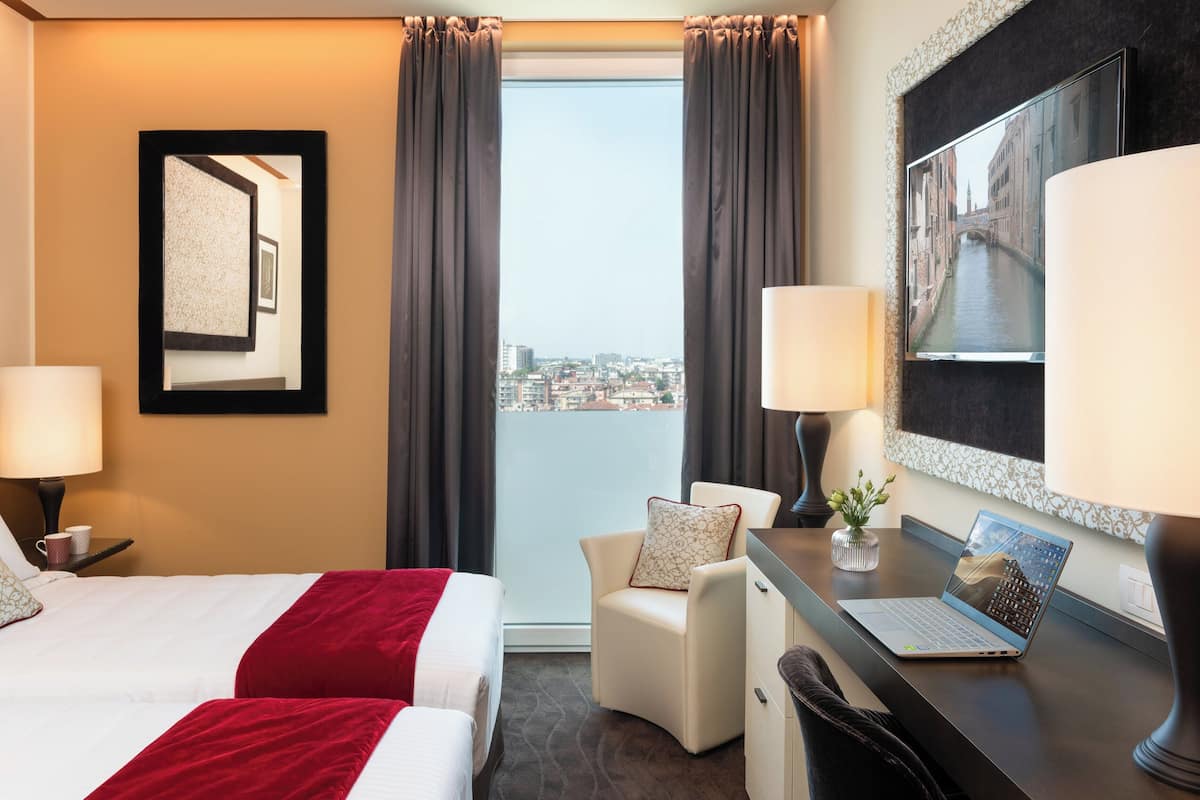 Una habitación de hotel con una cama, dos mesitas de noche, un escritorio con una computadora portátil, una silla, un ventanal con cortinas y vistas a la ciudad.