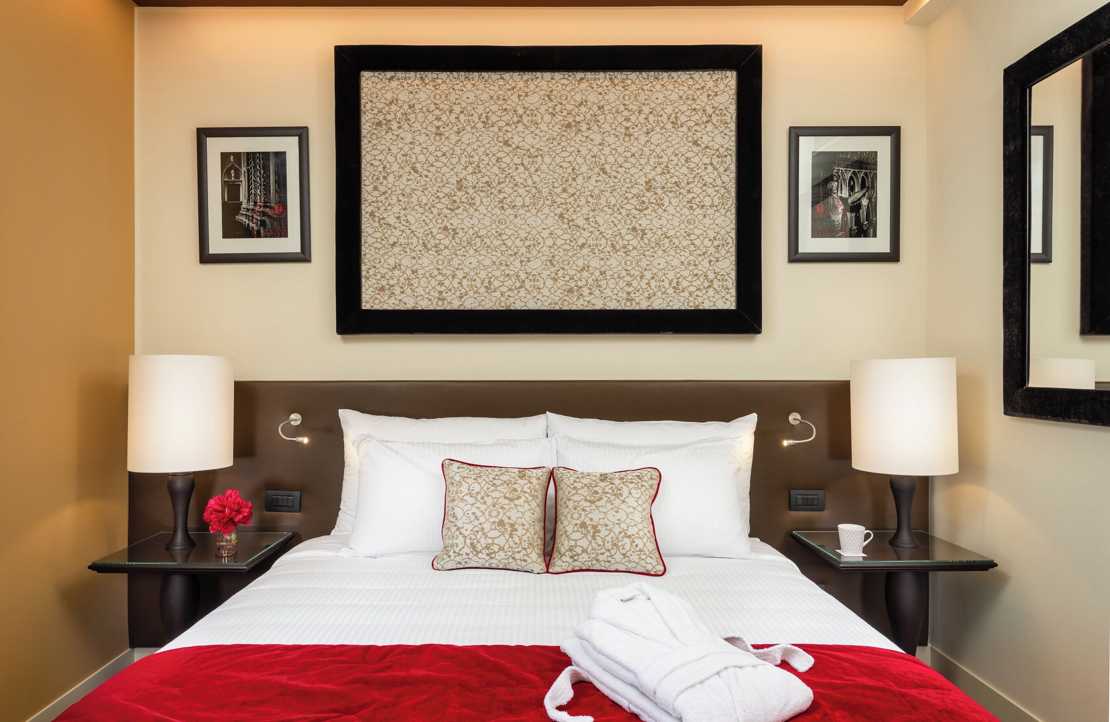 A neatly made bed with white linens and red accents, two bedside tables with lamps, and framed artwork on the walls.