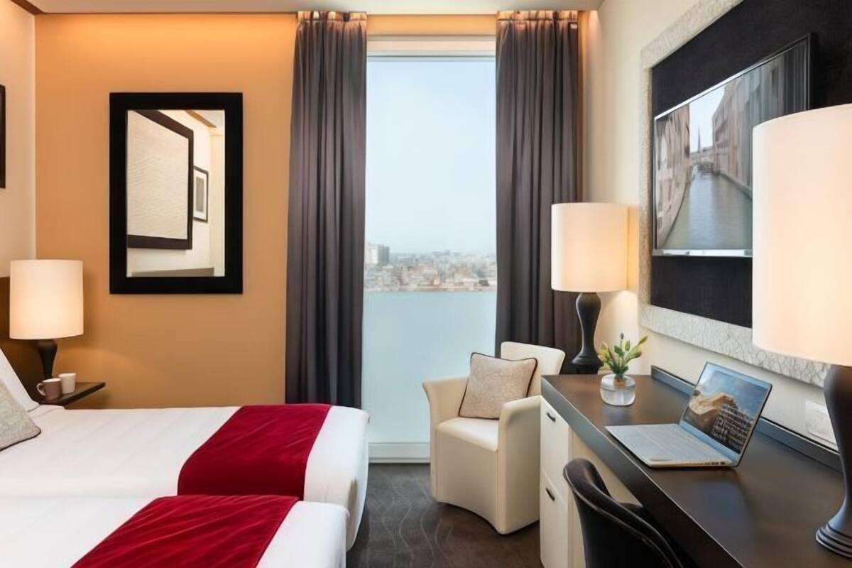 Habitación de hotel con cama, dos lámparas de noche, un escritorio con una computadora portátil, una silla y un ventanal con cortinas.