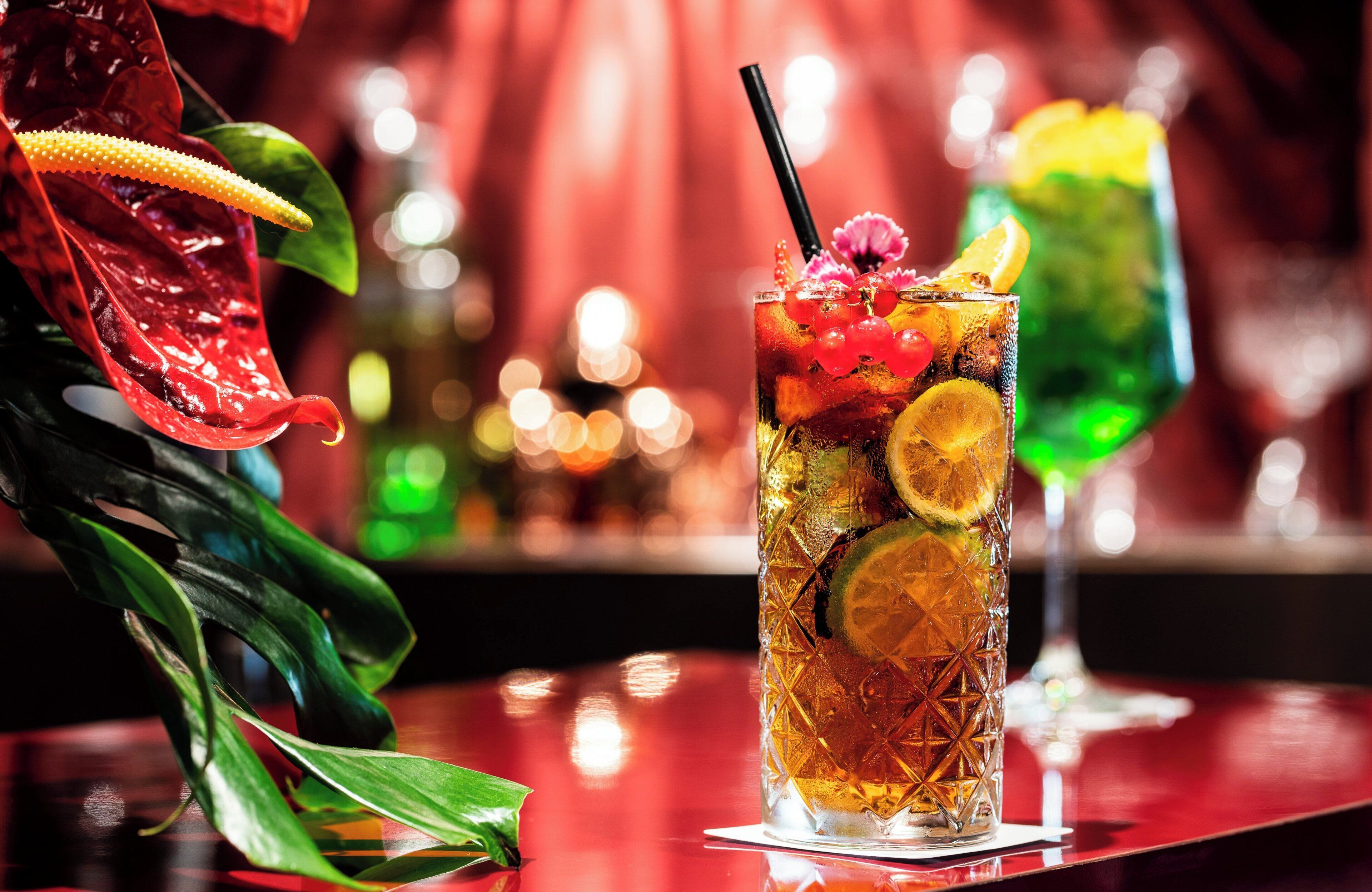 A tall glass with a straw, garnished with fruit and a flower, placed on a reflective surface with a blurred background.