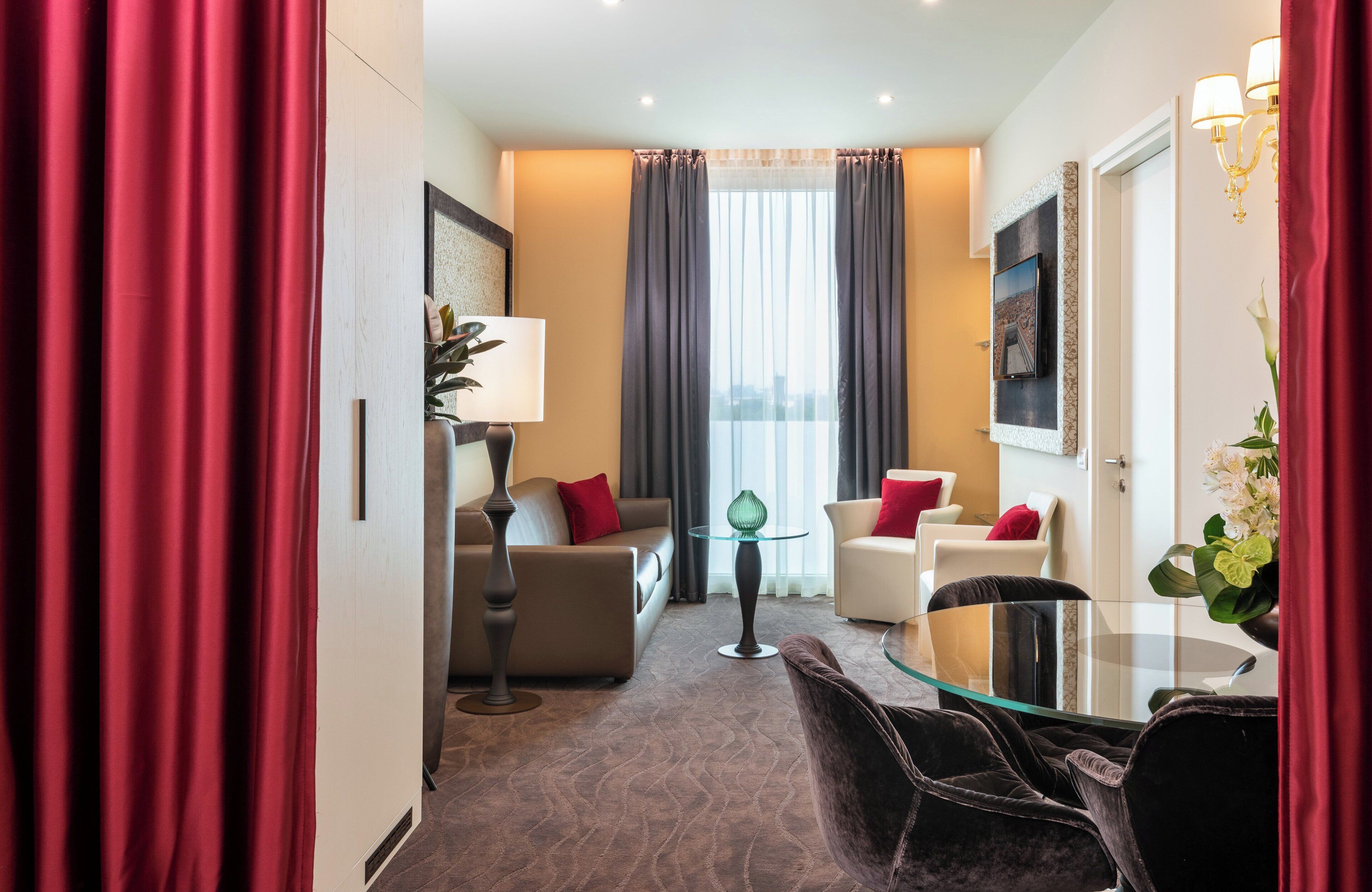 Een hotelkamer met een bank, fauteuils, een glazen tafel, een lamp en uitzicht op de stad door grote ramen.