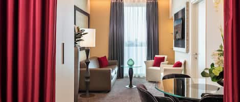 A hotel room with a sofa, armchairs, a glass table, a lamp, and a view of the city through large windows.