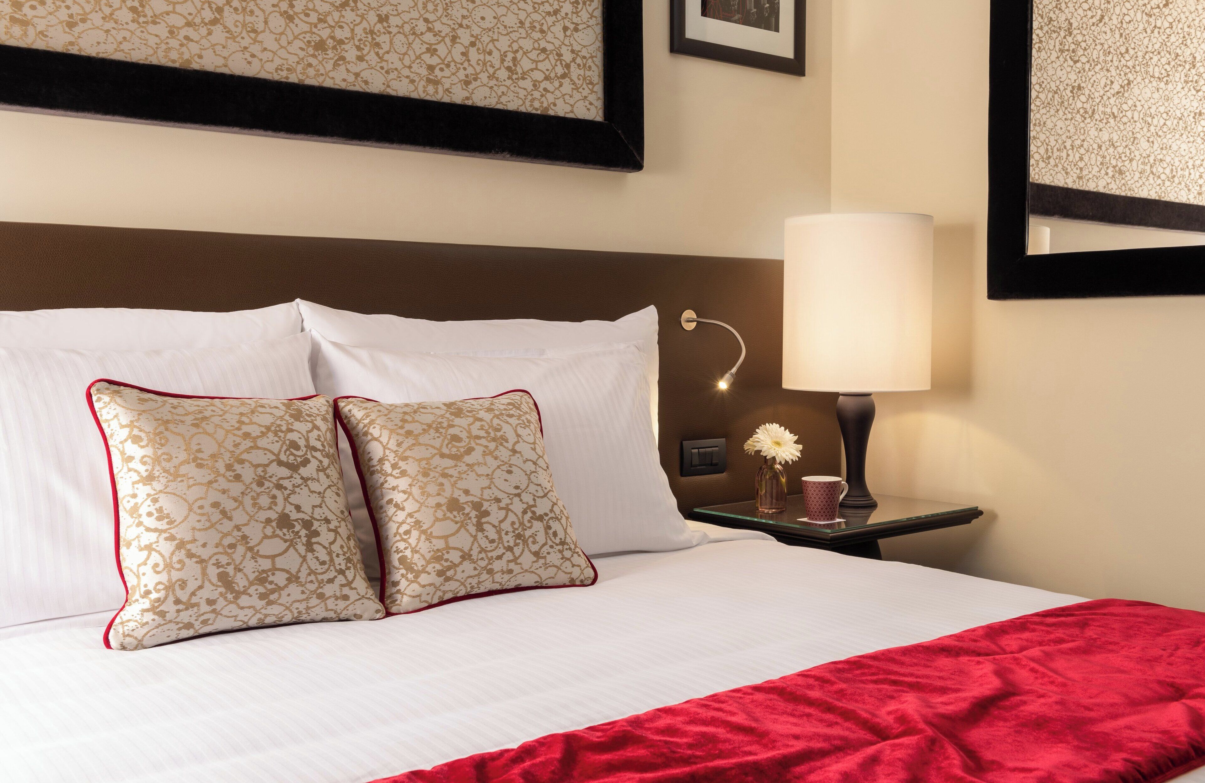 Una cama bien hecha con sábanas blancas y dos almohadas decorativas, una lámpara de noche y un pequeño jarrón con una flor.