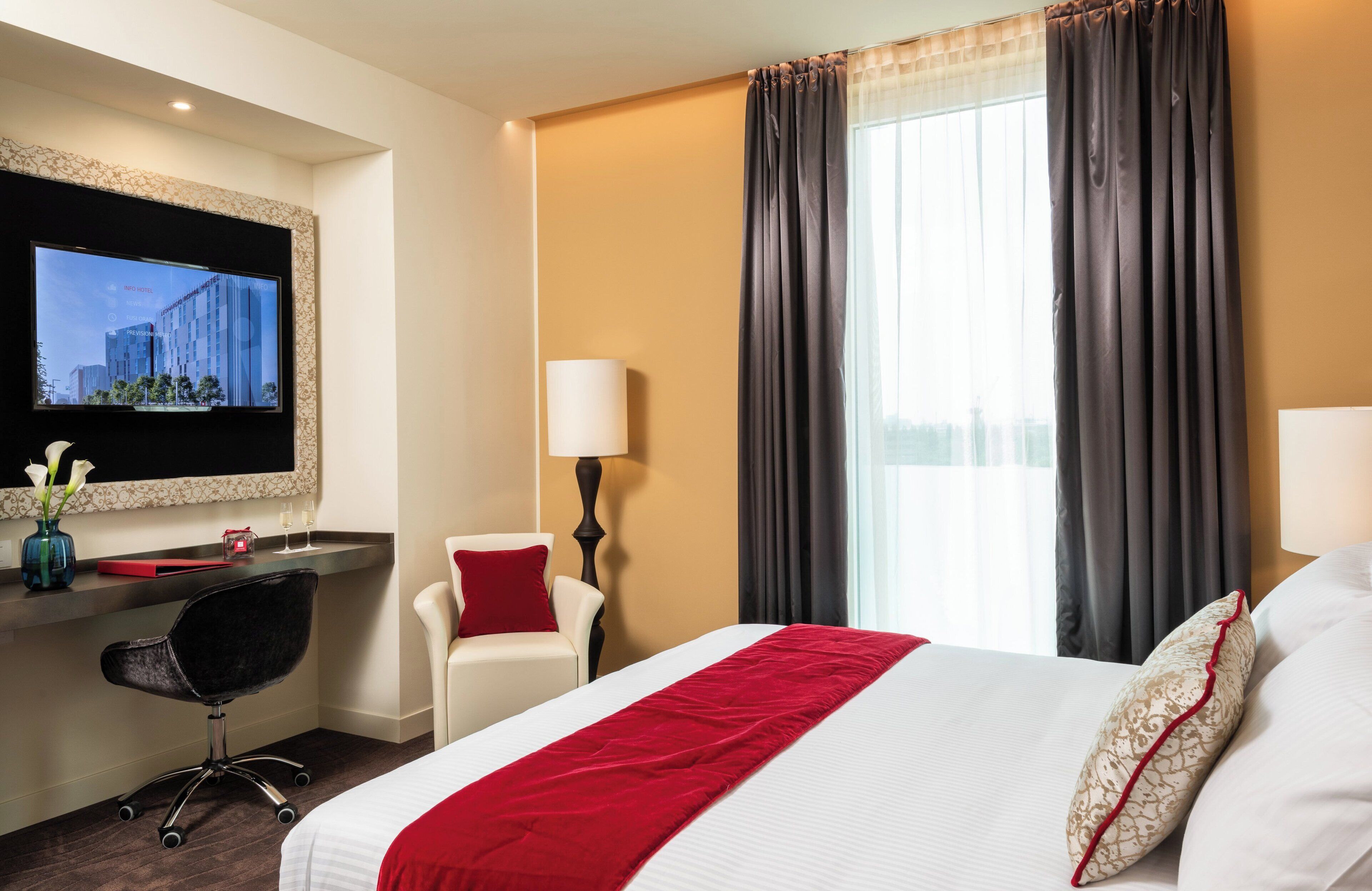 A hotel room with a bed, a desk with a chair, a TV, and a window with curtains.