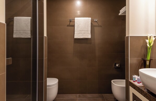 A modern bathroom with a glass shower enclosure, two white toilets, and a vanity with a large mirror and a vase of flowers.