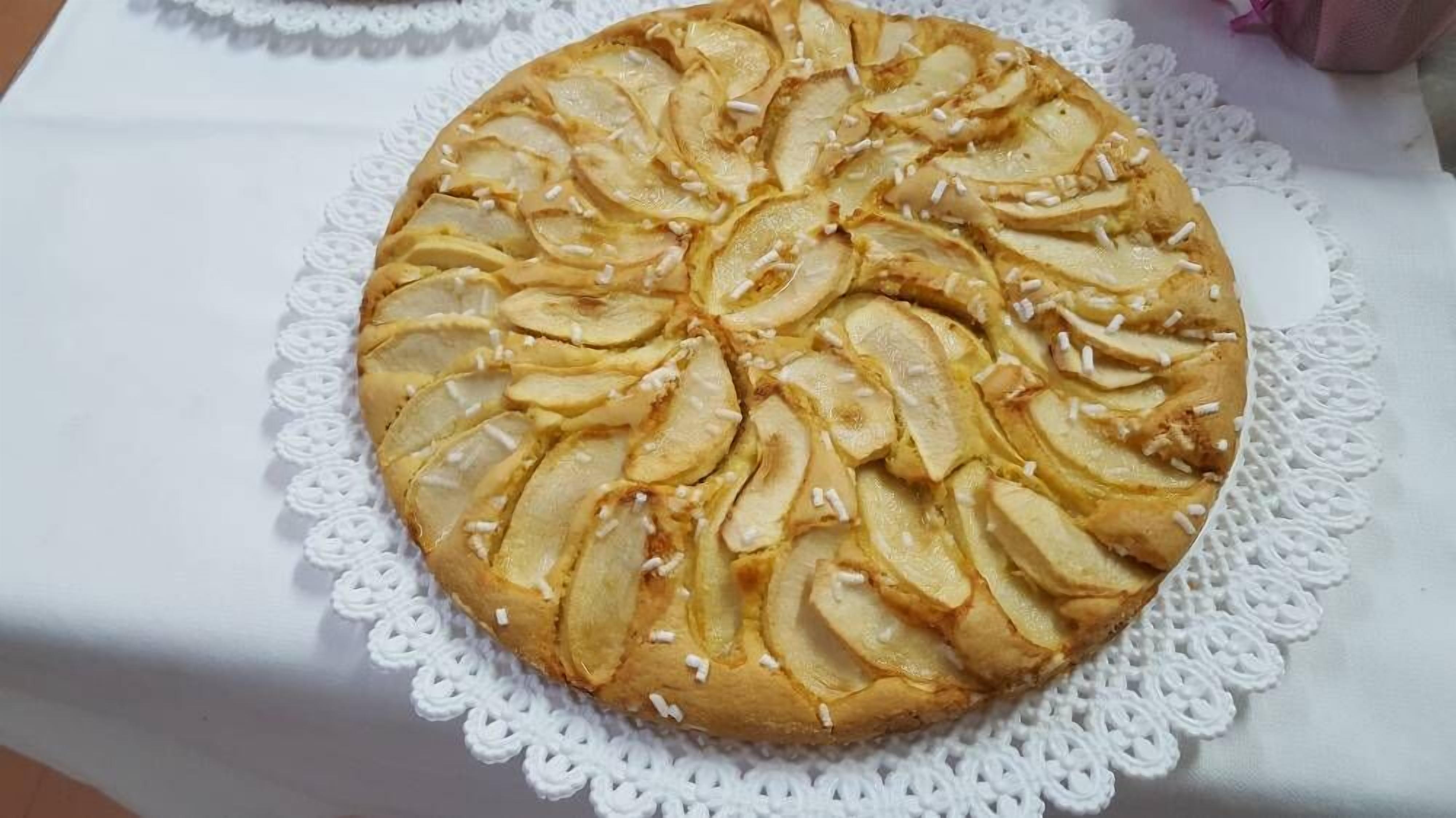 Una torta rotonda con fettine di mele sopra, su un tavolo bianco con un sottotavolo in pizzo.