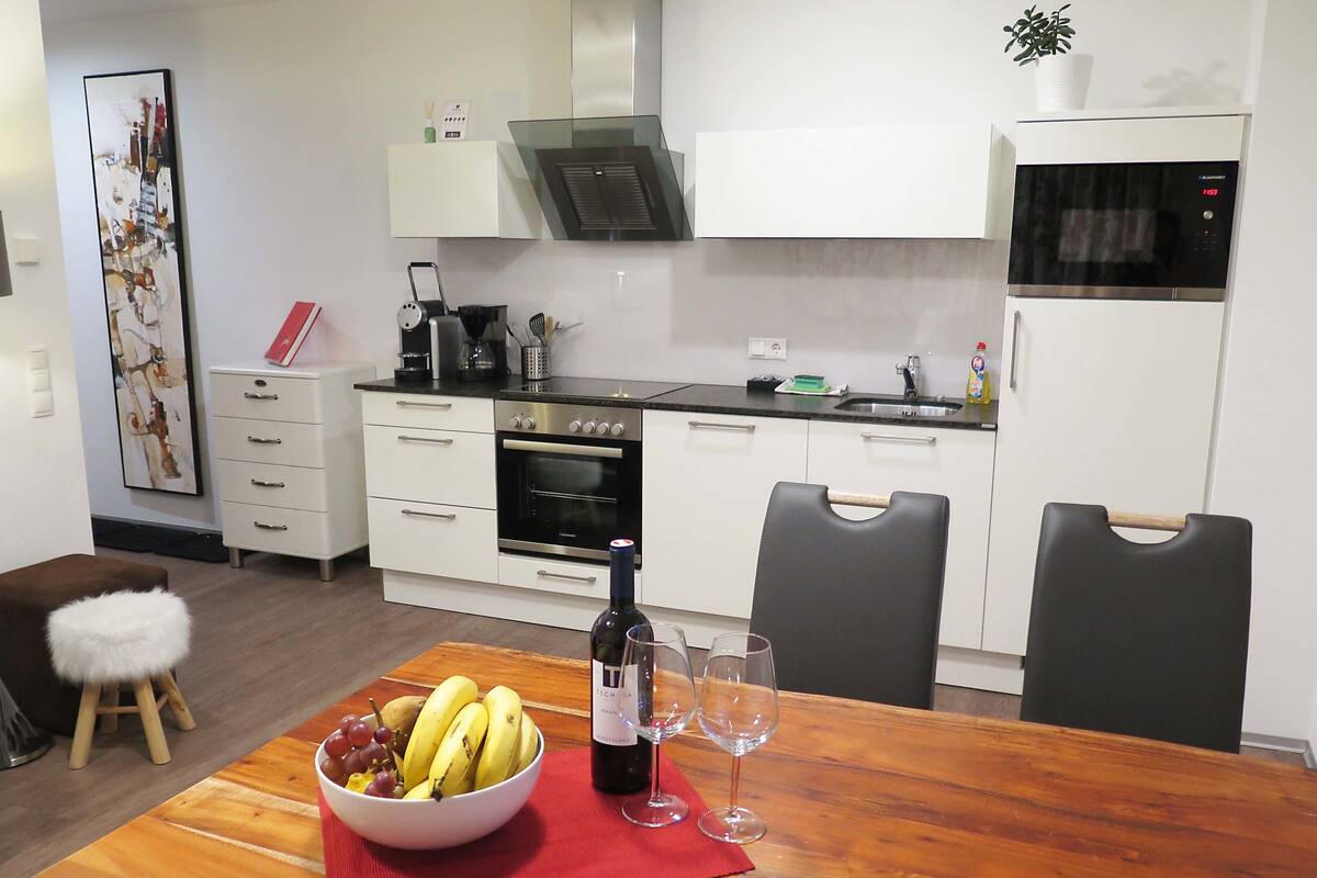 Een moderne keuken met witte kasten, een houten eettafel met een fruitschaal, wijnglazen en een fles wijn.